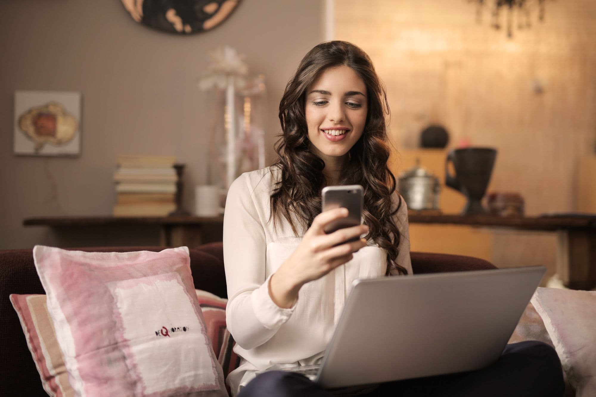 Freelancer kijkt glimlachend naar een melding op haar telefoon, laptop open op schoot in een gezellige woonkamer
