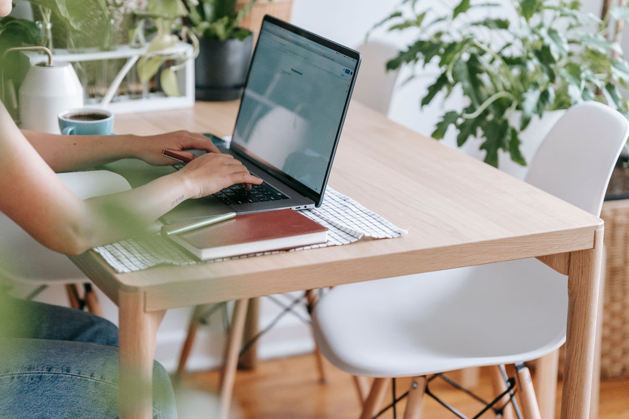 Freelancer typt op een laptop aan een houten tafel tussen kamerplanten, kopje koffie binnen handbereik